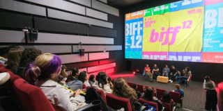 Imagen de varias personas en un escenario donde se ve una gran pantalla en la que se proyecta publicidad del BIFF 12.