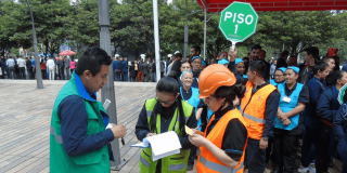 Personas participan en un ejercicio de evacuación en un espacio abierto. En primer plano, varios integrantes de brigadas de emergencia con chalecos reflectivos verdes y naranjas registran información en formularios. Detrás, un grupo organizado de participantes se reúne junto a una señal octagonal verde con el texto “PISO 1”.