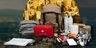 Kit de emergencias sobre una mesa con una mochila gris, botiquín rojo, linterna, pilas, radio portátil, alimentos no perecederos, botellas de agua y otros elementos recomendados para atender una emergencia.