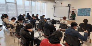Foto que muestra personas sentadas en escritorios en un salón de clases