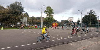 Persona con casco amarillo monta una bicicleta entre conos de señalización instalados en una pista durante Vacaciones Recreativas del IDRD con actividades de formación ciclista al aire libre.
