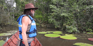 Persona con chaleco salvavidas observa un cuerpo de agua rodeado de vegetación en una zona selvática, donde se destaca la Victoria Colombiana con grandes hojas circulares flotando sobre la superficie.