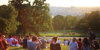 En un parque grande lleno de pasto hay varias personas sentadas compartiendo un picnic con la puesta del sol