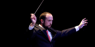 Un hombre joven, de cabello castaño y con algo de calvicie dirige una orquesta.