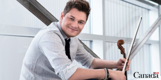 Hombre joven y blanco sostiene un violín mientras posa sentado y sonriente.