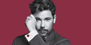 Hombre blanco de cabello castaño posa mirando a la cámara con una mano a un lado de su rostro.