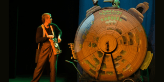 Imagen de un hombre frente a un relog de madera gigante alusivo a la obra de teatro 