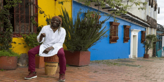 Imagen del maestro Nicoyembe sentado a las afueras de una casa antigua de color azul rey y amarillo