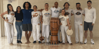 Imagen de seis bailarines de capoeira en escenario acompañados de instrumentos músicales