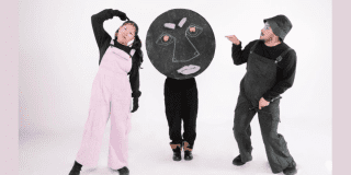 Imagen de tres personajes, uno viste un overol rosado y el otro verde oscuro, el tercer personaje lleva un gran círculo negro con un rostro dibujado en tiza que cubre su cuerpo.