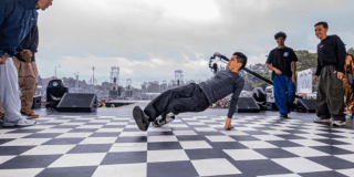 Grupo de jóvenes bailando en una pista con apariencia de ajedrez, de fondo se ve mucha gente observándolos
