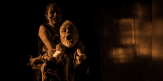 Imagen de la puesta en escena donde se ve a una mujer que ahorca por la espalda a un hombre de barba que está sendado en un silla