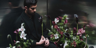 Imagen del cantante sentado dentro de un asensor, rodeado de muchas flores.