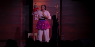 Imagen de los protagonistas de la obra: una mujer joven vestida de balnco con un delantal y un títere femenino con vestido rojo.