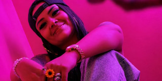 Imagen de 'Angelik DK' sentada con gorra y anillos en un fondo rosado.
