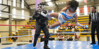 Una competidora de lucha libre realiza una patada aérea contra un luchador enmascarado dentro del cuadrilátero, mientras un árbitro y espectadores observan el combate.