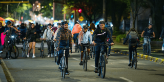 Ciclistas y peatones recorren una avenida de Bogotá durante la Ciclovía Nocturna, con grupos de personas en bicicleta, alumbrado público, árboles y actividad recreativa en horario nocturno.