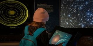 Imagen de una niña con maleta y gorro de espaldas interactuando con pantalla en el Planetario de Bogotá