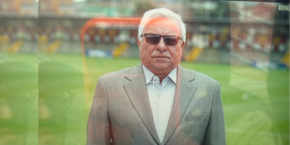 Imagen de un hombre mayor que usa gafas oscuras, al fondo se ve una cancha de fútbol.