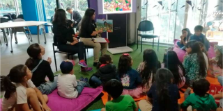 Iamgen de varios niños y niñas sentados en colchonetas atendiendo la charla de la tallerista que indica en un libro.