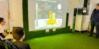 Imagen de dos niños sentados observando una presentación en una pantalla gigante, al lado hay un hombre adulto hablando. 