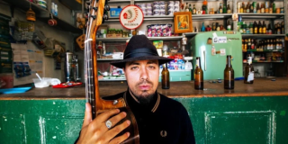 Imagen del cantante en una tienda de barrio con una guitarra en la mano, de fondo se ven varias botellas de cerveza.