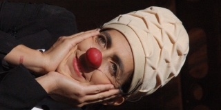 Imagen de la cara de una mujer caracterisada como payasa, con una nariz redonda roja y un sombrero blanco.