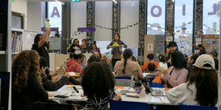 Actividad cultural en una biblioteca pública de Bogotá, con varias personas sentadas alrededor de mesas mientras observan una presentación en un escenario al frente. En el espacio se ven estanterías, elementos decorativos y letras colgantes.