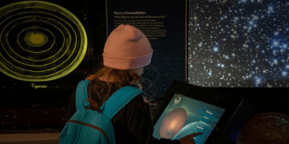 Imagen de una niña con maleta y gorro de espaldas interactuando con pantalla en el Planetario de Bogotá
