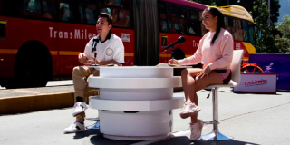 Imagen de un hombre y una mujer, presentadores de Cicloviva sentados en una vía de Bogotá, de fondo se ve un bus de Transmilenio rojo.