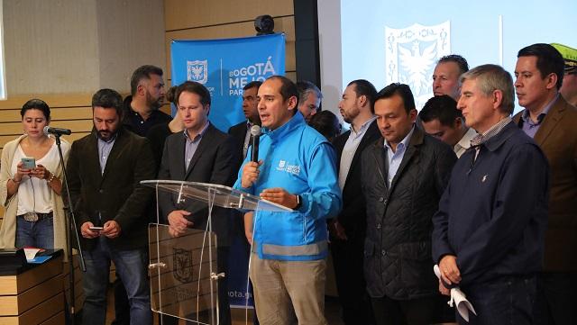 Varias personas reunidas en una tarima, un hombre de chaqueta azul dirige una conferencia con el microfono en la mano
