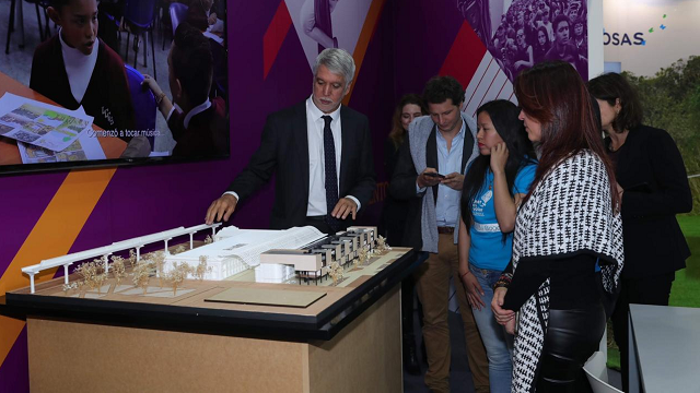 El Alcalde Enrique Peñalosa expone una obra en maqueta que esta exhibida en el stan de la Alcaldía de Bogotá a varias personas que lo observan 