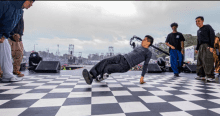 Grupo de jóvenes bailando en una pista con apariencia de ajedrez, de fondo se ve mucha gente observándolos