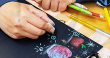 Imagen de una mano con una tiza sobre cartelera negra con dibujos y colores