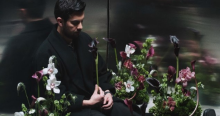 Imagen del cantante sentado dentro de un asensor, rodeado de muchas flores.