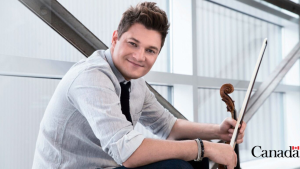 Hombre joven y blanco sostiene un violín mientras posa sentado y sonriente.