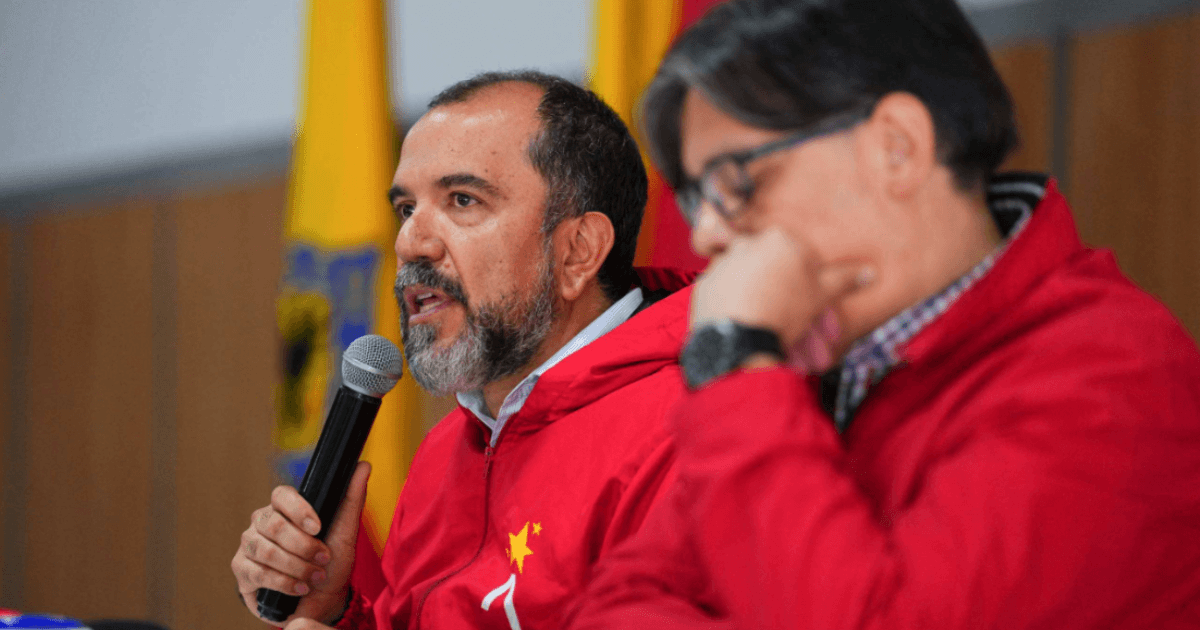 Primer plano de una mesa de reunión institucional. El director del IDRD, Daniel García Cañón, con chaqueta roja sostiene un micrófono y habla, mientras otra persona, también con chaqueta roja, aparece desenfocada en primer plano durante rueda de prensa para explicar situación de la gramilla del estadio El Campín de Bogotá.