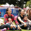Imagen de mamás con niños en sus piernas sentadas en el pasto en un parque prestando atención a alguien, de fondo se ve la naturaleza del parque