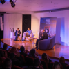 Panel de conversación en auditorio con cuatro personas sentadas en el escenario frente a público asistente, iluminación azul y morada, plantas decorativas y banner al fondo.