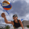 Jugadora de voleibol playa golpea un balón Mikasa durante un partido sobre arena, con gafas deportivas, malla de voleibol y palmeras visibles bajo un cielo parcialmente nublado.
