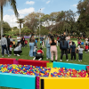 Personas adultas y menores participan en actividades recreativas sobre una zona verde de parque; en primer plano hay piscinas secas con pelotas de colores y al fondo se observan conos, árboles y grupos reunidos al aire libre.