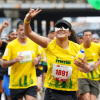 Corredores participan en la Media Maratón de Bogotá mientras avanzan por una avenida de la ciudad; en primer plano una atleta con camiseta amarilla y número 1891 saluda al público durante la competencia.