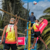 Partido de voleibol playa con dos deportistas junto a la red durante competencia al aire libre, con palmeras y juez en silla de arbitraje.