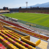 Vista interior del estadio El Campín en Bogotá con graderías amarillas y rojas, cancha de fútbol en óptimas condiciones y montañas al fondo.