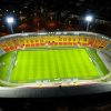 Vista aérea nocturna del estadio El Campín en Bogotá, iluminado por reflectores, con gramilla, graderías multicolores y entorno urbano visible alrededor.