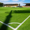 Cancha de fútbol del estadio de Techo con césped renovado, líneas demarcadas, arcos y graderías vacías bajo cielo parcialmente despejado.