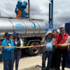 Foto que muestra un grupo de personas sonriendo sosteniendo una cinta y un carrotanque en el fondo