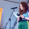 Imagen de una mujer joven en una tarima leyendo un libro de poesia, al lado inzquierdo se ve una pancarta del concurso Reverso.