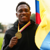 Ronal Longa sosteniendo una medalla dorada colgada al cuello y una bandera de Colombia. Al fondo se observa una pista o campo deportivo con vegetación desenfocada.