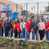 Grupo de personas con chaquetas rojas y ropa casual de pie junto a un jardín recién sembrado, frente a una estructura metálica y edificaciones del sector. En primer plano se observan plantas jóvenes y áreas con tierra.